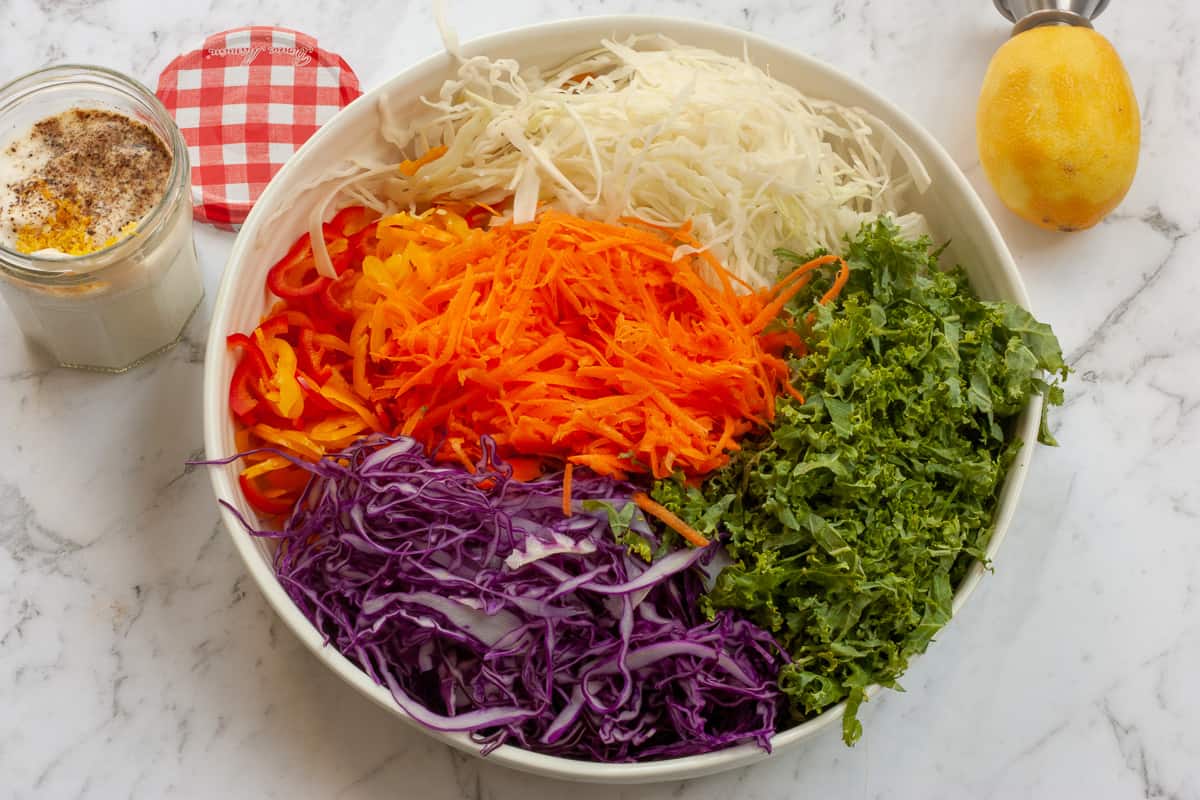 Piles of shredded vegetables in a a white bowl. The salad dressing is in a jam jar to the side.