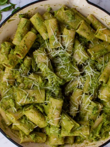 Big red circular pan containing rigatoni pasta with a green pesto, sliced broccoli florets and grated parmesan.