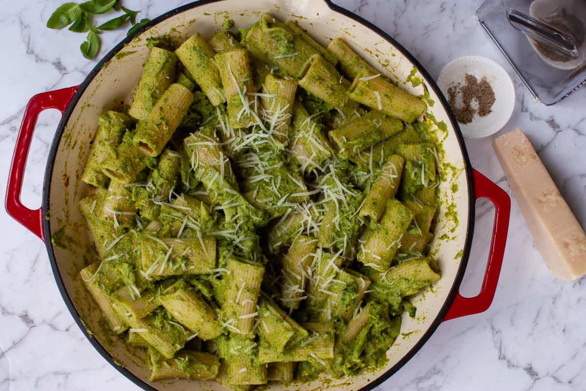 Big red circular pan containing rigatoni pasta with a green pesto, sliced broccoli florets and grated parmesan.