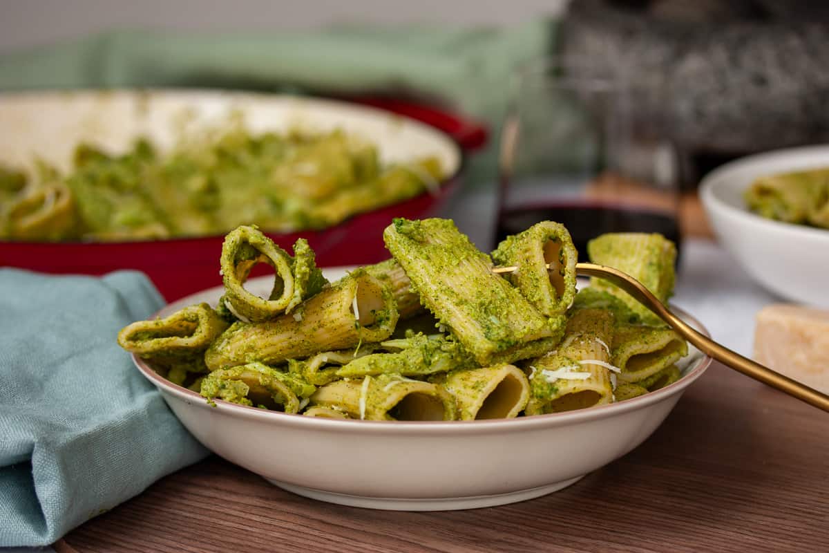 Bowl of heaped rigatoni covered in green pesto. A couple of pasta pieces speared with a gold fork.