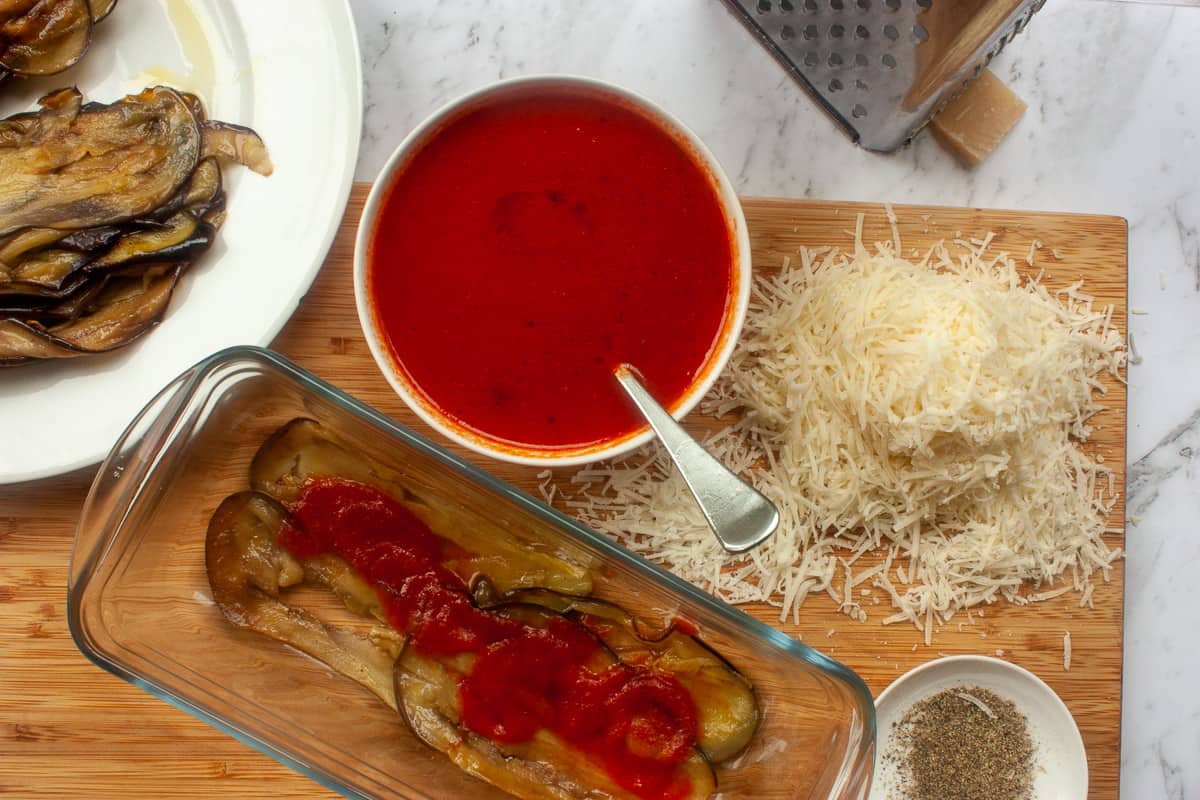 Layering aubergine, tomato sauce and grated cheese in a glass loaf pan. All the ingredients are around the loaf pan.