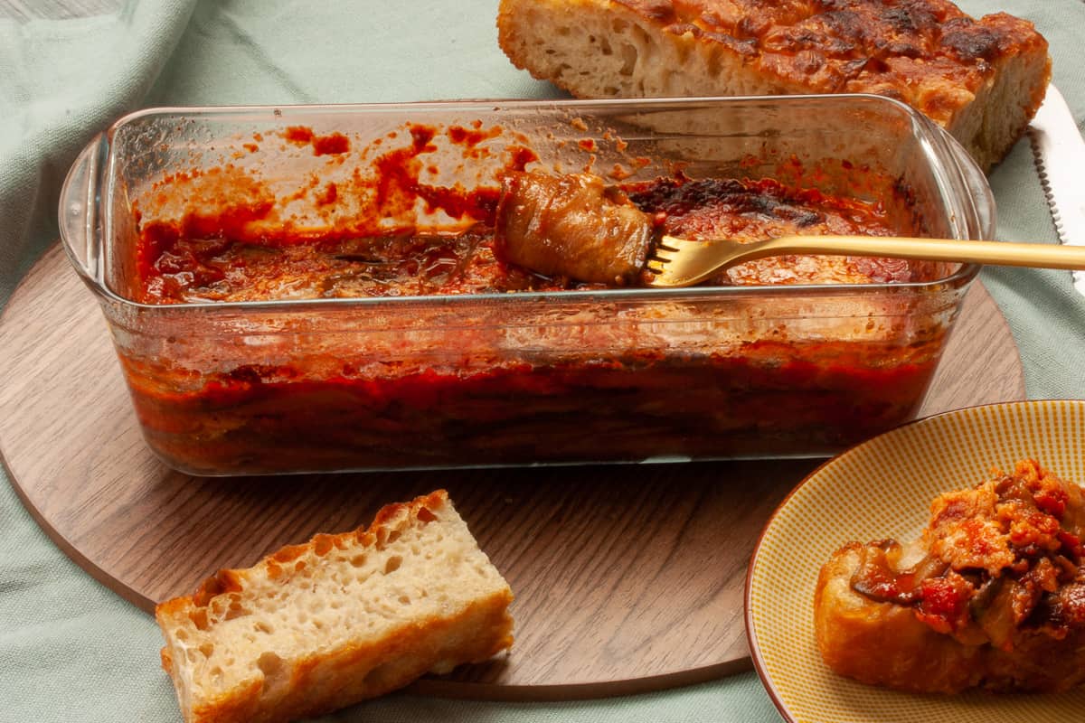 Aubergine parmigiana slice twirled around a fork. Some already on a piece of bread, with the loaf of bread in the background.