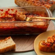 Aubergine parmigiana slice twirled around a fork. Some already on a piece of bread, with the loaf of bread in the background.