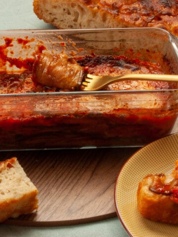 Aubergine parmigiana slice twirled around a fork. Some already on a piece of bread, with the loaf of bread in the background.