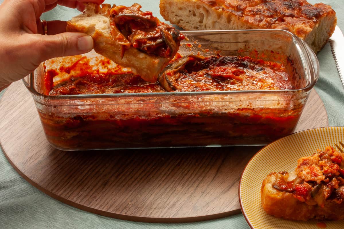 A hand holding a big chunk of bread loaded with aubergine parmigiana.