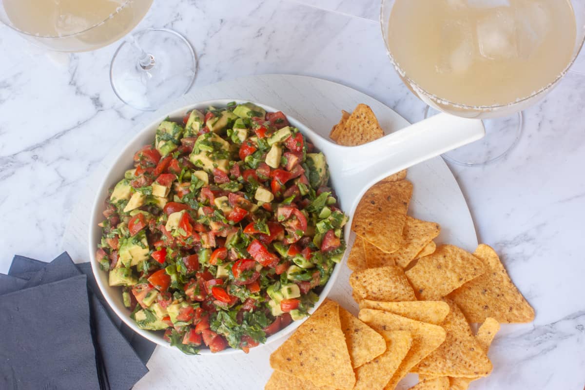 Avocado corn chip topper in a white bowl surrounded by triangular corn chips and a couple of margarita cocktails.