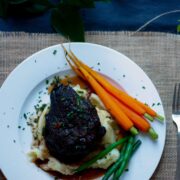 Overhead view of a whole beef cheek atop a bed of mashed potato. with baby carrots and steamed green beans on a white plate.