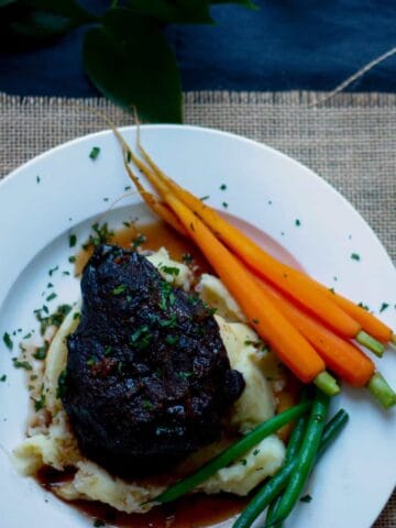 Overhead view of a whole beef cheek atop a bed of mashed potato. with baby carrots and steamed green beans on a white plate.