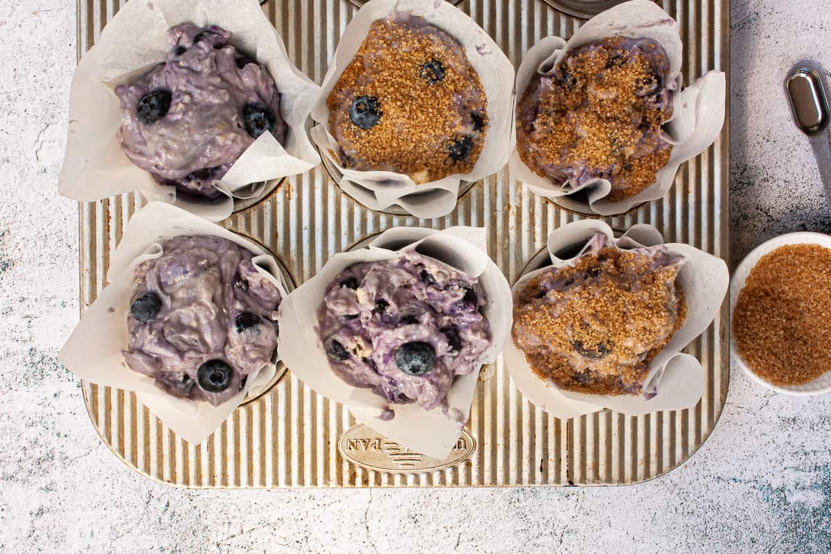 Blueberry muffin batter in paper cases. Half are sprinkled with coarse brown sugar.