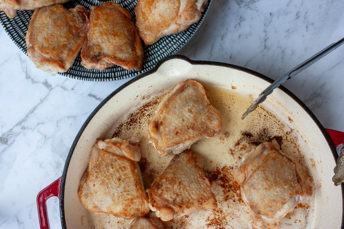 Chicken pieces being browned in a pan, some browned pieces on a plate nearby.