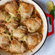 Top view of bronzed chicken pieces in a red baking dish with thinly sliced lemon slices nestled amongst them.