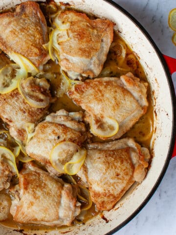 Top view of bronzed chicken pieces in a red baking dish with thinly sliced lemon slices nestled amongst them.