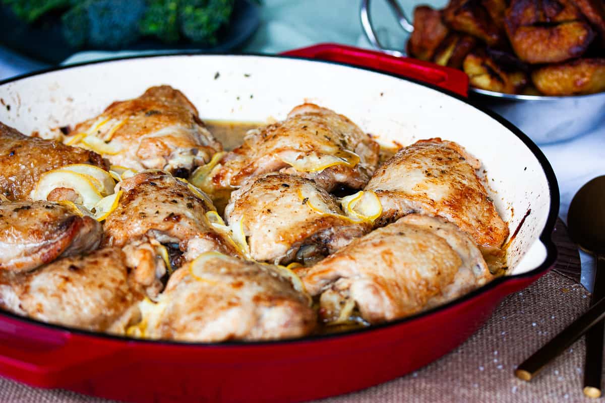 Bronzed chicken pieces in a red baking dish with thinly sliced lemon slices nestled amongst them.