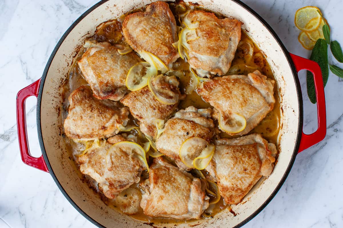 Top view of bronzed chicken pieces in a red baking dish with thinly sliced lemon slices nestled amongst them.