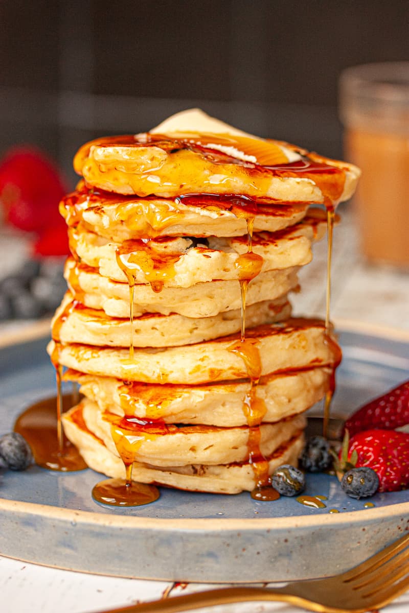 A tall stack of buttermilk pancakes. There is some melted butter at the top and maple syrup drizzled all over.