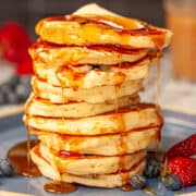 A tall stack of buttermilk pancakes. There is some melted butter at the top and maple syrup drizzled all over.