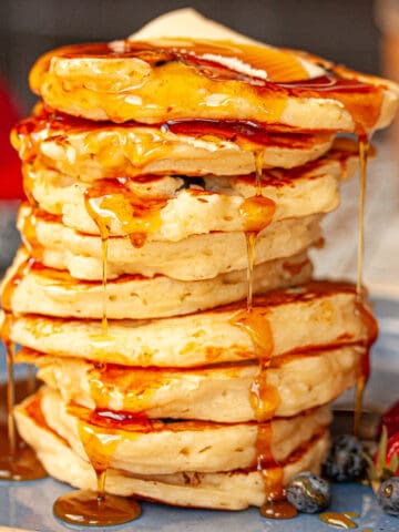 A tall stack of buttermilk pancakes. There is some melted butter at the top and maple syrup drizzled all over.