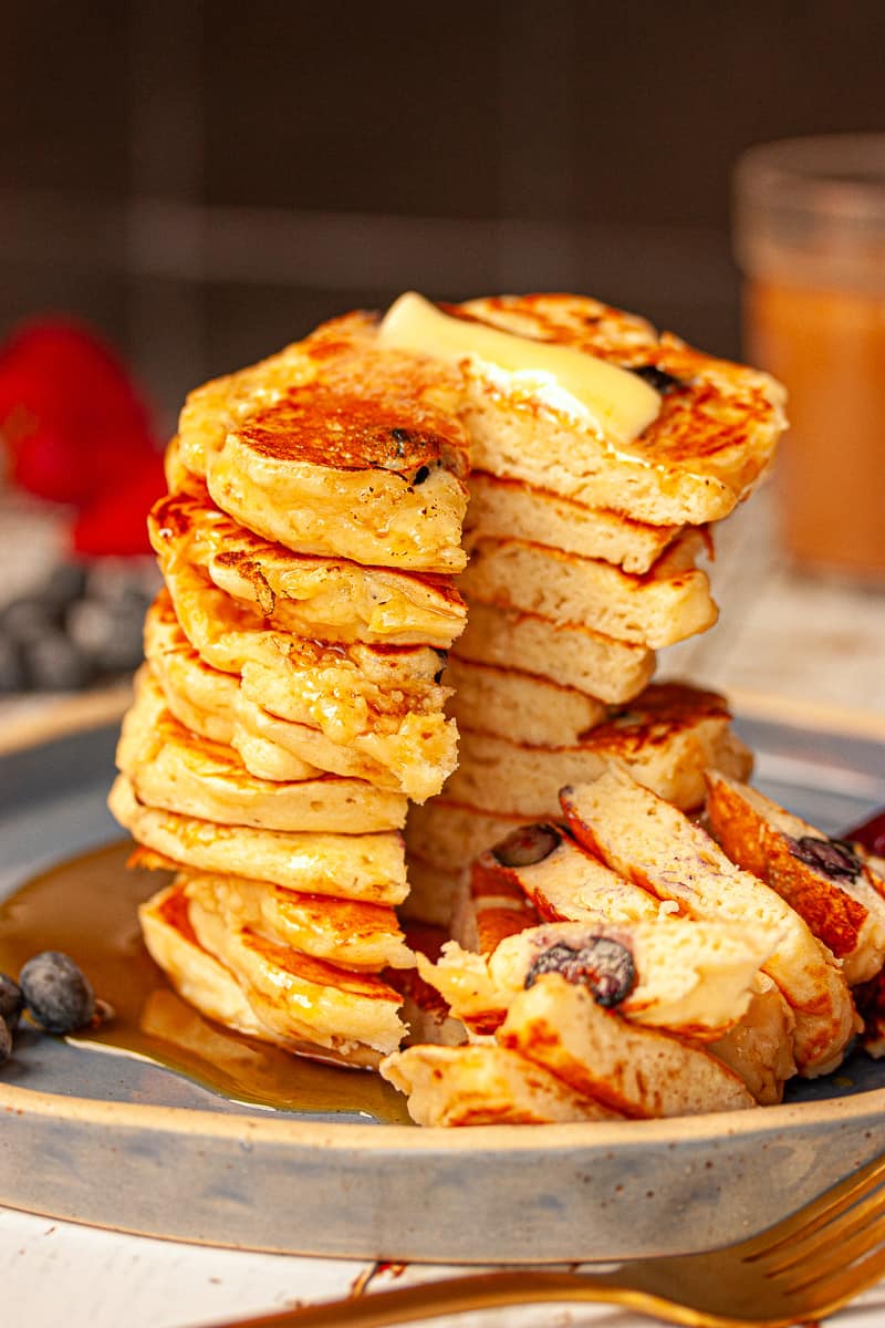 A tall stack of buttermilk pancakes with a wedge cut through them. There is some melted butter at the top and maple syrup drizzled all over.