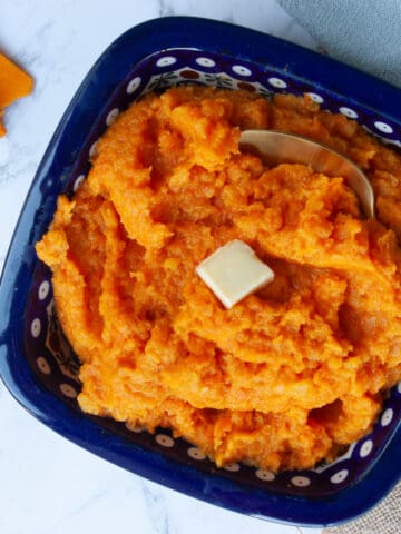 Square blue bowl containing orange mashed veg with a square of melting butter on the top.