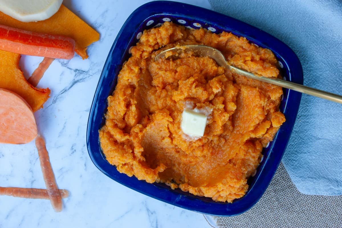 Square blue bowl containing orange mashed veg with a square of melting butter on the top.