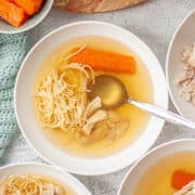 Looking down into a white bowl of clear yellow broth with fine noodles, carrot and shredded chicken.