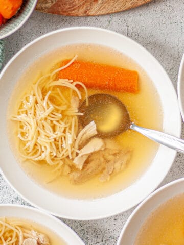 Looking down into a white bowl of clear yellow broth with fine noodles, carrot and shredded chicken.