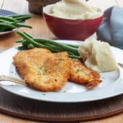 A white plate with a pair of chicken schnitzel scattered with chopped parsley with some mashed potato and green beans.