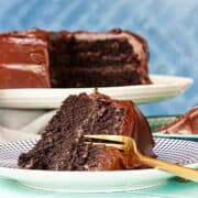 Big slice of chocolate cake with a gold fork inserted ready to take a bite. The whole cake is in the background.