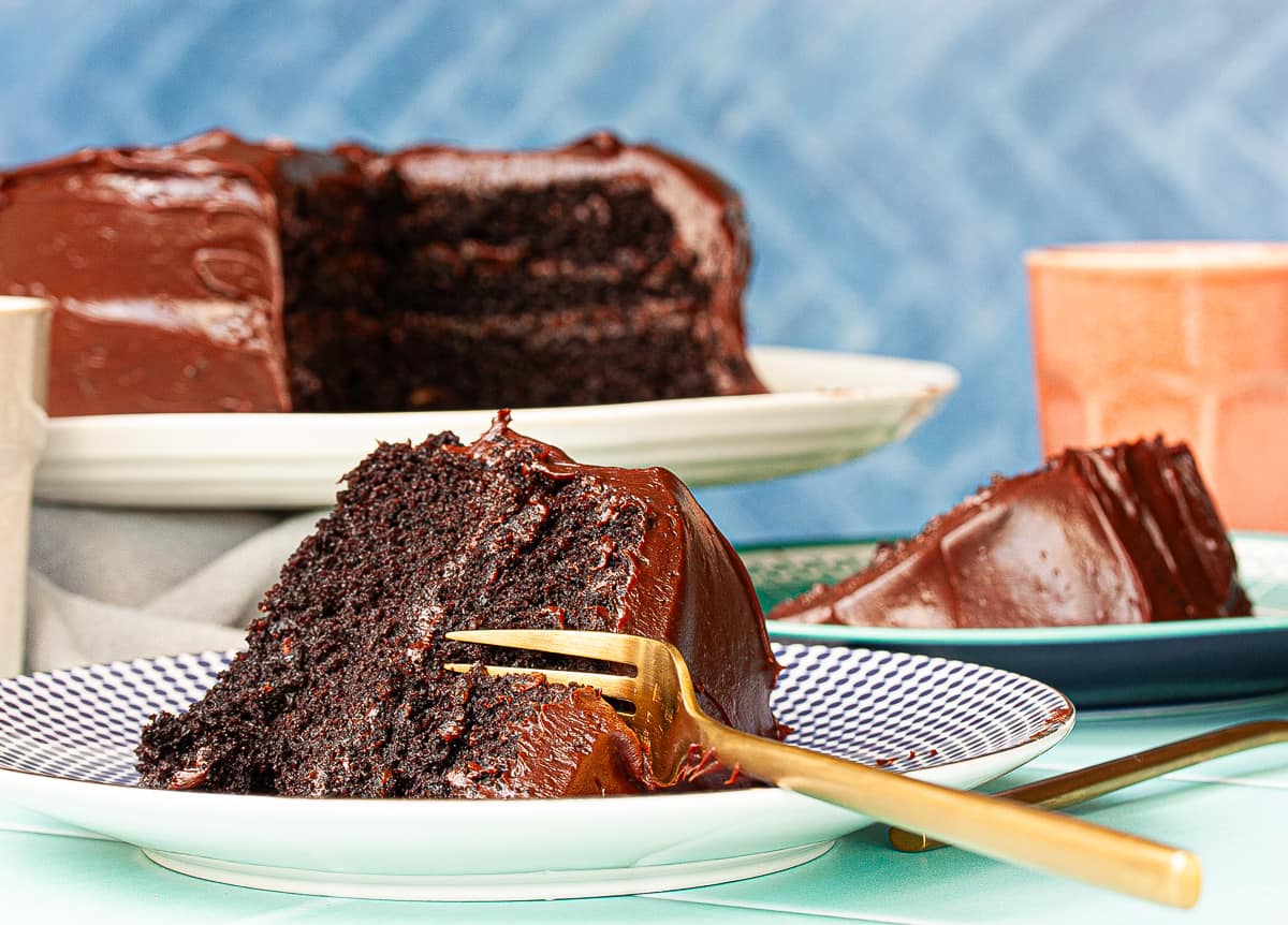 Big slice of chocolate cake with a gold fork inserted ready to take a bite. The whole cake is in the background.