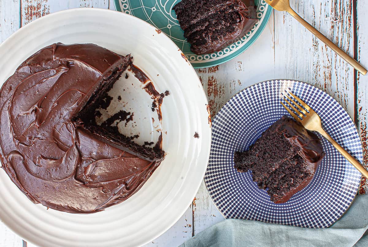 Top view of a richly frosted chocolate cake on a white platter. Two slices cut from it are on side plates.