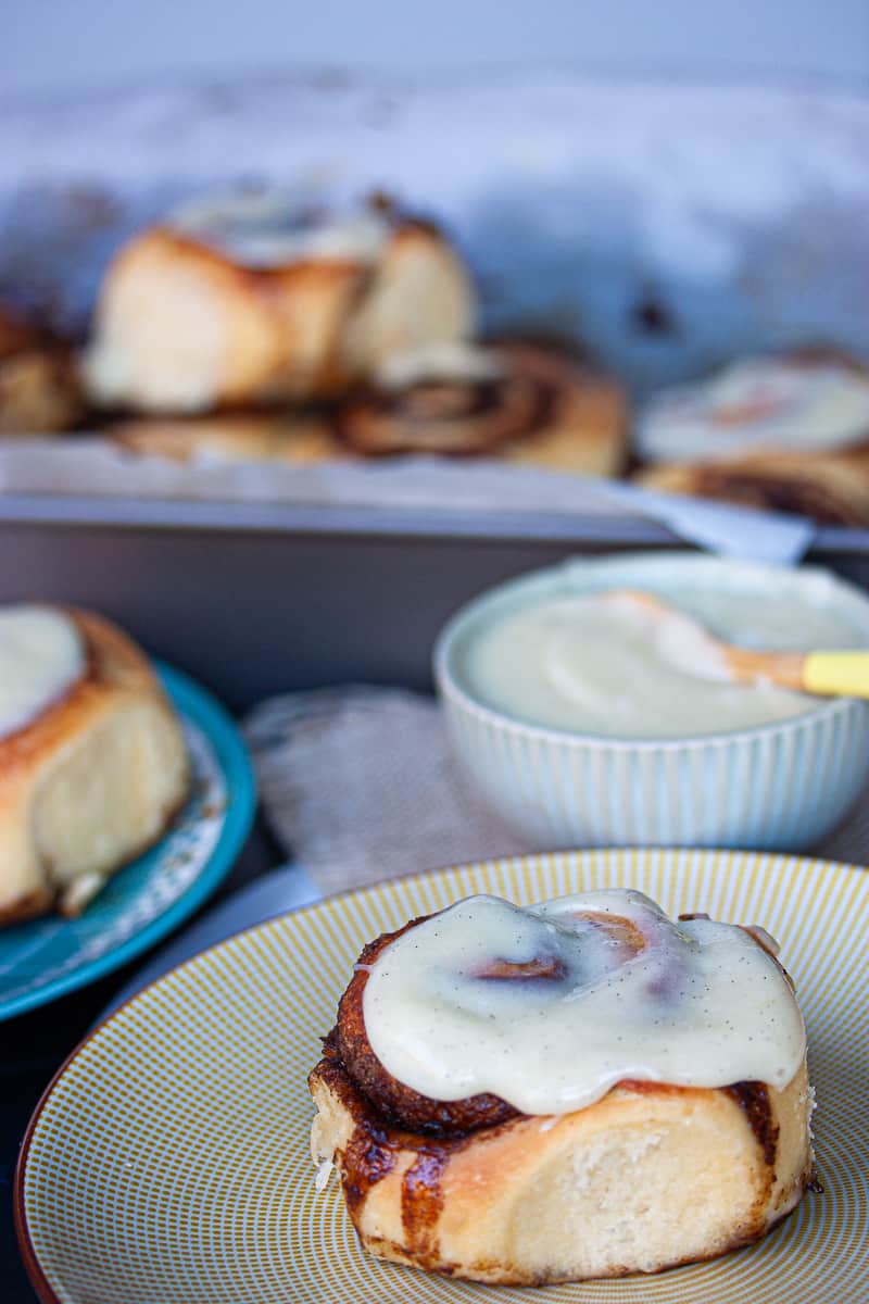 Close up of an iced cinnamon bun on a plate. The tray of cinnamon buns is in the background.