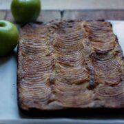 Close up of apple cake removed from baking dish and set on baking paper. Whole green apples and cinnamon sticks to the side.