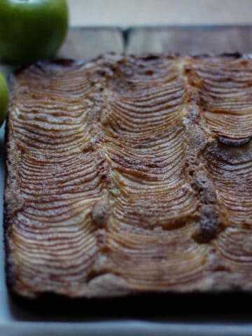 Close up of apple cake removed from baking dish and set on baking paper. Whole green apples and cinnamon sticks to the side.