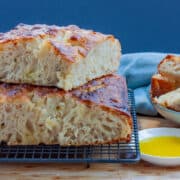 Two stacked pieces of focaccia bread showing all the bubbles inside along side oil dipping bowl and a bowl of bread chunks.