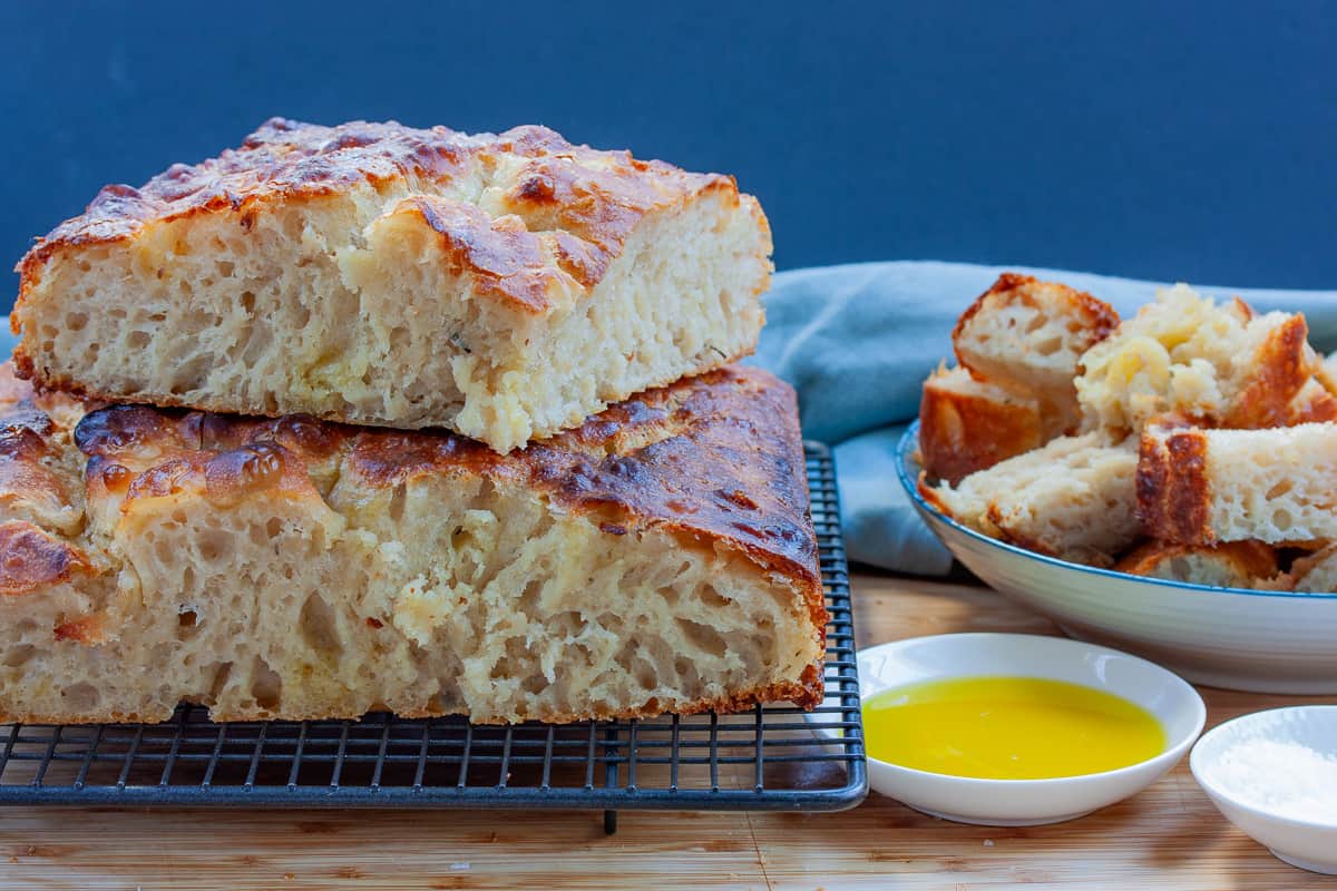 Two stacked pieces of focaccia bread showing all the bubbles inside along side oil dipping bowl and a bowl of bread chunks.