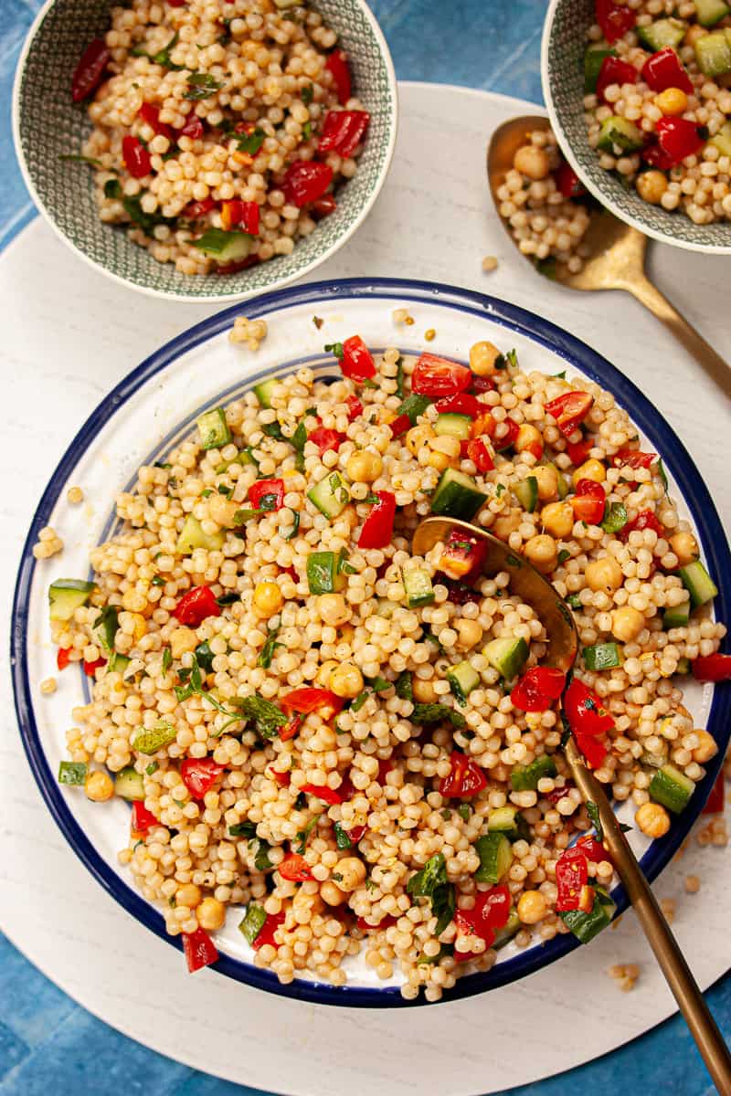 Colourful giant couscous salad in a large bowl with two smaller filled serving bowls.