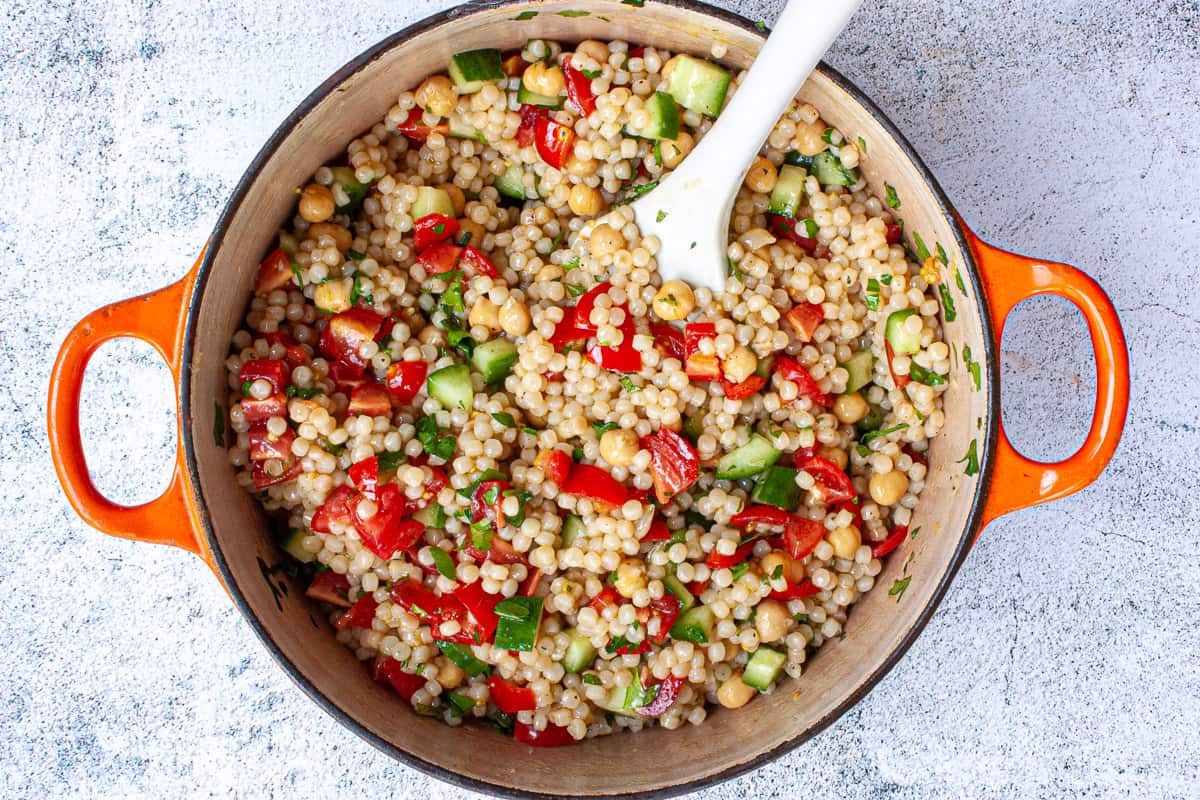 Colourful giant couscous salad in a pot with orange handles.