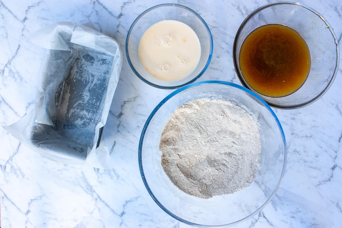 Three glass bowls containing batter ingredients set next to a lined loaf pan.