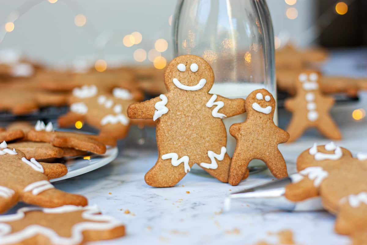 Happy gingerbread people propped up against a small glass bottle of milk. Other biscuits around them.