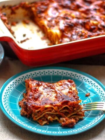 Generous portion of lasagna on a small blue plate. There's another serving in the background and the whole lasagna with two pieces missing.