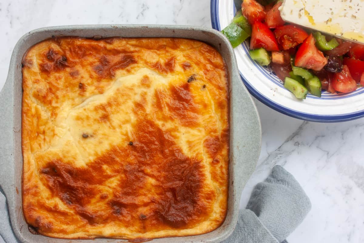 Whole baked moussaka in a grey square baking dish. There is a Greek salad next to it.