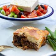 Square portion of moussaka on a plate with green beans. There is Greek salad in the background.