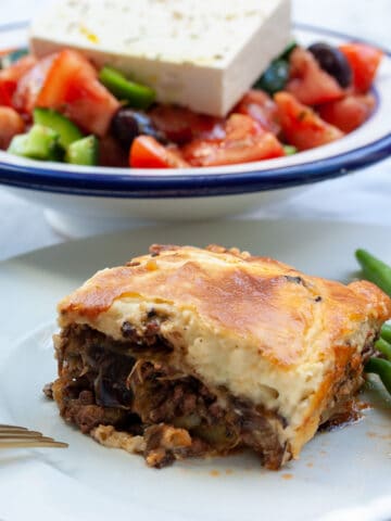 Square portion of moussaka on a plate with green beans. There is Greek salad in the background.