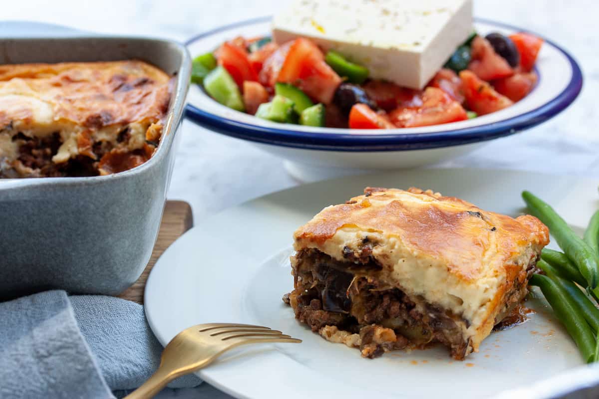 Square portion of moussaka on a plate with green beans. There is Greek salad in the background.