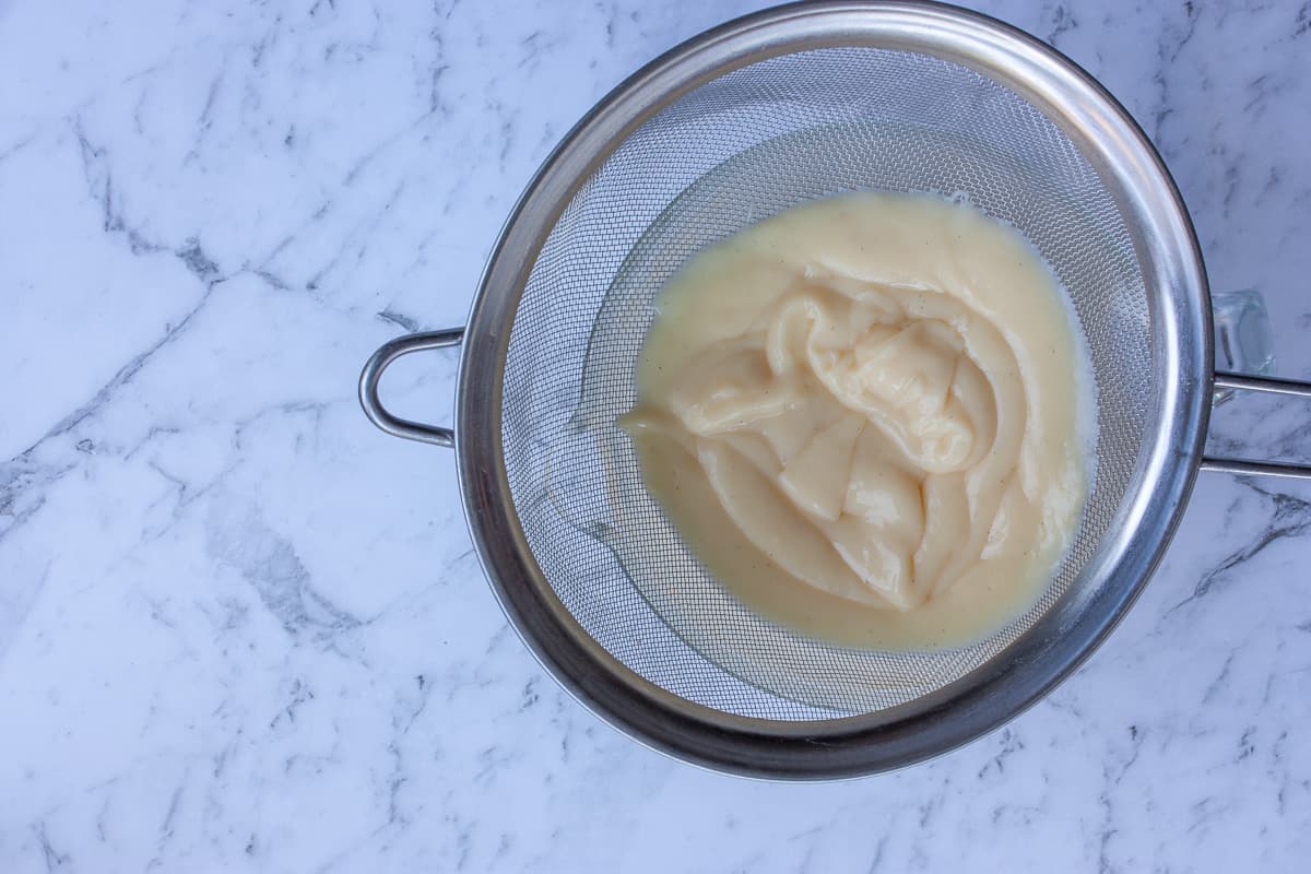 Cooked pastry cream being sieved into a glass bowl.