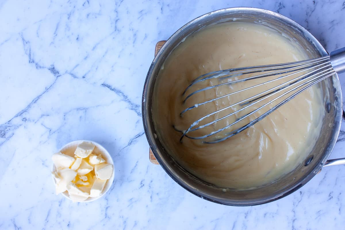 Hand whisk resting in thick custard in a saucepan. Cubed butter in a small white dish alongside.