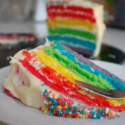 Close-up of a slice of rainbow cake on a white plate. The whole cake is in the background.