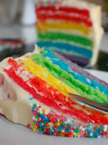 Close-up of a slice of rainbow cake on a white plate. The whole cake is in the background.