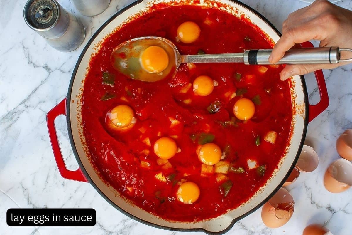 Eggs freshly cracked into shallow wells in the shakshouka sauce. A large shallow serving spoon is being used to do this.