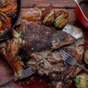 Partially shredded lamb on a wooden board surrounded by sides - ratatouille tian, potato stacks and gravy.